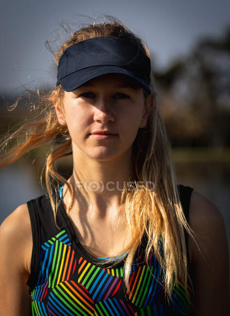 Retrato close-up de uma remadora caucasiana de uma equipe de remo com cabelo loiro longo, vestindo uma viseira, de pé ao sol, olhando para a câmera, com o rio no fundo — Fotografia de Stock