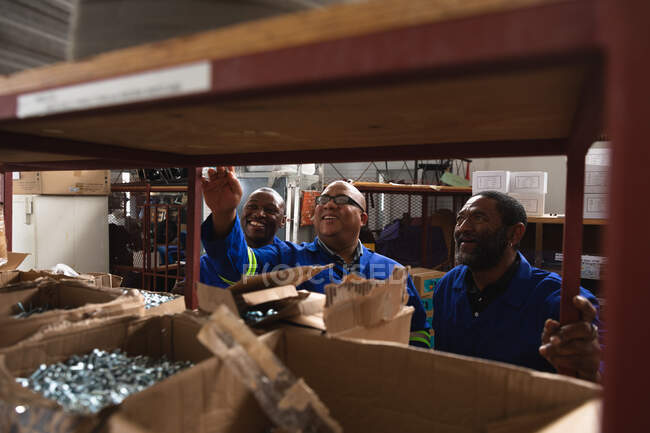 Троє кавказьких і афроамериканських чоловіків на складі на заводі роблять інвалідні візки, стоять і перевіряють деталі на полицях. — стокове фото