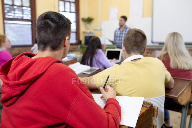 Rückansicht einer multiethnischen Gruppe von Teenagern in einem Klassenzimmer der High School, die am Schreibtisch sitzt und ihrem männlichen kaukasischen Lehrer zuhört, der vor einer Klasse steht — Stockfoto