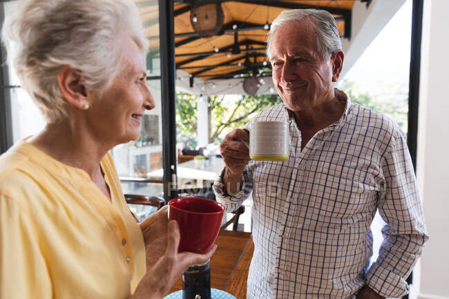 Старшая кавказская пара дома на кухне, разговаривает, улыбается и пьет кофе вместе в солнечный день, пара изолируется во время пандемии коронавируса — стоковое фото