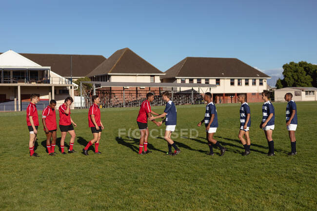Vista laterale di due squadre maschili multietniche adolescenti di giocatori di rugby che indossano la loro striscia di squadra, si salutano sul campo di gioco, si stringono la mano prima dell'inizio della partita — Foto stock