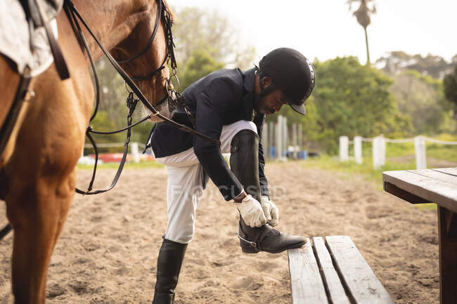 Side view of a smartly dressed African American man putting riding boots on before dressage horse riding during a sunny day, with a chestnut horse standing by him on a paddock. — Stock Photo