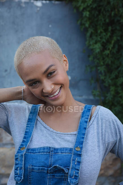 Portrait of mixed race alternative woman with short blonde hair wearing denim dungarees, out and about in the city on a sunny day, looking to camera and smiling. Urban independent woman on the go. — Stock Photo
