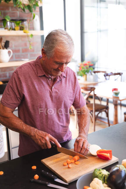 Feliz jubilado mayor hombre caucásico en casa en la cocina en un día soleado, de pie en la encimera picando verduras en una tabla de cortar y sonriendo, auto aislante durante coronavirus covid19 pandemia - foto de stock