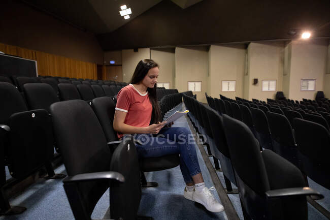Vista laterale di un'adolescente caucasica in un teatro vuoto del liceo, seduta nell'auditorium che si prepara per uno spettacolo, con una sceneggiatura e linee di apprendimento — Foto stock