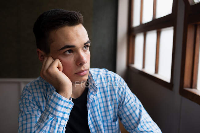 Side view close up of a teenage Caucasian boy in a school classroom sitting at desk, concentrating, looking out of the window, leaning his head on his hand — Stock Photo