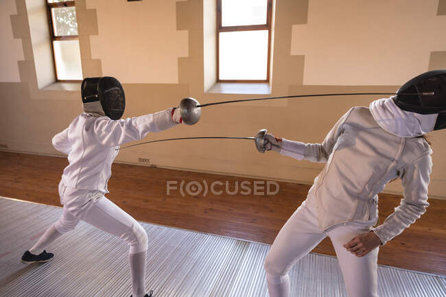 Caucasian and African American sportswomen wearing protective fencing outfits during a fencing training session, lunging at each other with their epees. Fencers training at gym. — Stock Photo