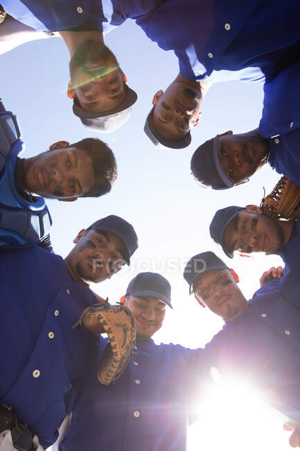Visão de baixo ângulo de uma equipe multi-étnica de jogadores masculinos de beisebol, preparando-se antes de um jogo, motivando uns aos outros em um aconchego, olhando para uma câmera, em um dia ensolarado — Fotografia de Stock