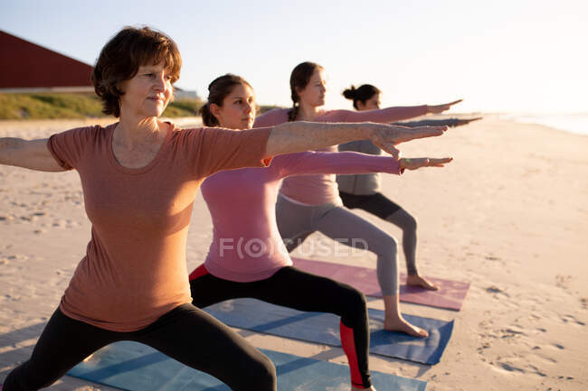Vista lateral de un grupo multiétnico de amigas disfrutando de hacer ejercicio en una playa en un día soleado, practicando yoga de pie en la asana guerrera. - foto de stock