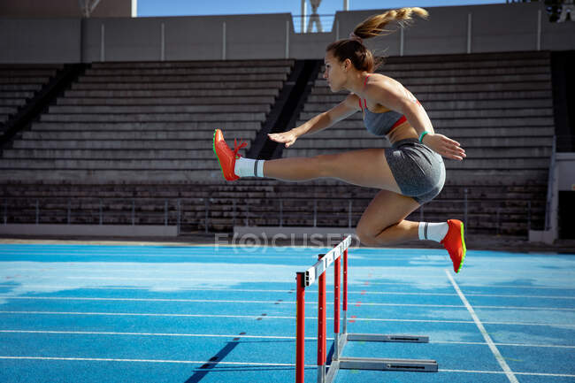 Вид сбоку кавказской спортсменки, практикующей на спортивном стадионе, бегущей и бегущей с барьерами. Лёгкая атлетика на стадионе. — стоковое фото