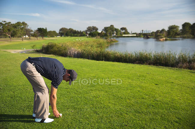 Вид сбоку на кавказца на поле для гольфа в солнечный день с голубым небом, помещающего мячик для гольфа на траву — стоковое фото