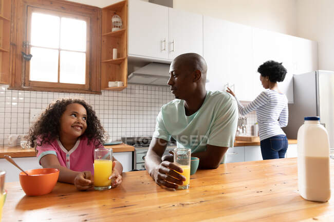 Vista frontal de una joven afroamericana y su padre en casa en la cocina por la mañana, sentados en la isla de la cocina con vasos de jugo de naranja hablando y sonriendo, la madre de pie en el fondo - foto de stock