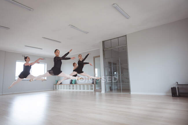 Um grupo de dançarinas de balé atraentes do sexo feminino caucasianas em roupas pretas praticando durante uma aula de balé em um estúdio brilhante, dançando e pulando no ar em uníssono . — Fotografia de Stock