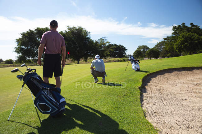 Vista trasera de dos hombres caucásicos en un campo de golf en un día soleado con cielo azul, de pie preparándose para el juego - foto de stock