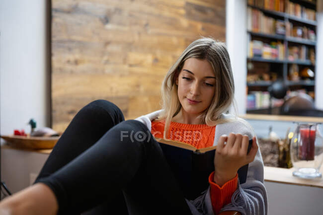 Vista frontal de cerca de una joven mujer caucásica, disfrutando del tiempo en casa, sentada en la sala de estar, leyendo un libro - foto de stock