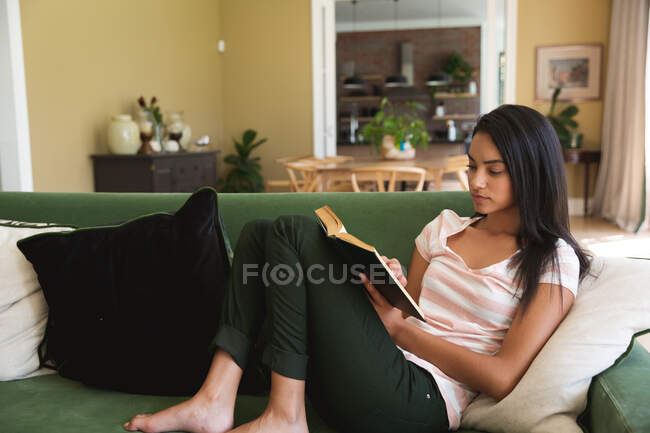 Mixed race woman spending time at home, reading a book in living room. Self isolating and social distancing in quarantine lockdown during coronavirus covid 19 epidemic. — Stock Photo
