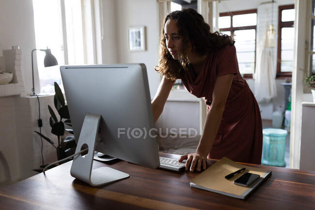 Mujer caucásica pasando tiempo en casa, de pie junto a su escritorio y trabajando con su computadora. Distanciamiento social y autoaislamiento en cuarentena. - foto de stock