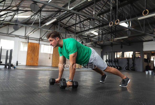 Side view of an athletic Caucasian man wearing sports clothes cross training at a gym, weight training with dumbbells, leaning in a press up position holding a dumbbell in each hand — Stock Photo
