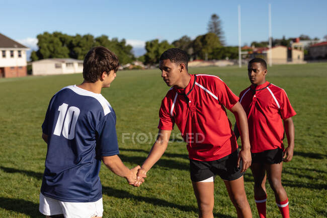 Vue latérale de trois joueurs de rugby masculins multi-ethniques adolescents d'équipes opposées portant leur bande d'équipe, se saluant sur le terrain de jeu et se serrant la main avant un match — Photo de stock