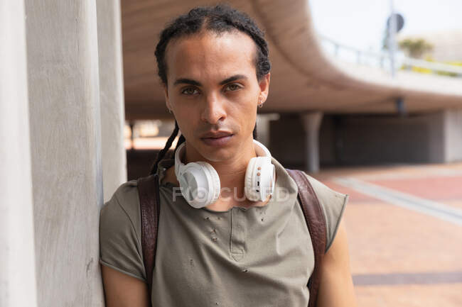 Ritratto di un uomo di razza mista con lunghi dreadlocks in giro per la città in una giornata di sole, in piedi in strada vicino a un ponte e guardando dritto in una macchina fotografica — Foto stock