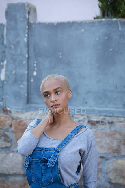 Mixed race alternative woman with short blonde hair wearing denim dungarees out and about in the city on a sunny day, standing by a wall and looking away. Urban independent woman on the go. — Stock Photo