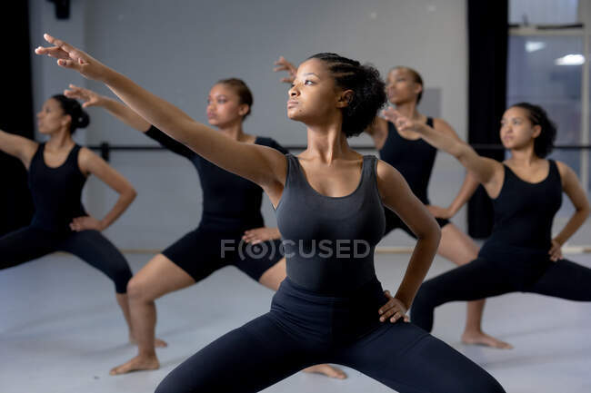 Vista frontal de un grupo multiétnico de bailarinas modernas en forma con trajes negros que practican una rutina de baile durante una clase de baile en un estudio brillante, extendiendo sus brazos. - foto de stock