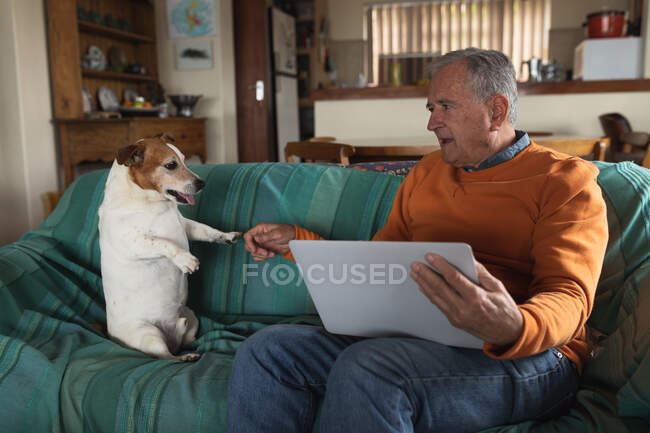 Vista frontal de un hombre caucásico mayor que se relaja en casa en su sala de estar, sentado en el sofá hablando con su perro mascota y utilizando una computadora portátil - foto de stock