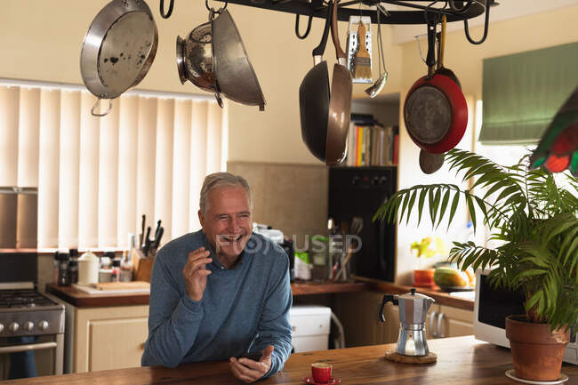 Vista frontal de un hombre caucásico mayor relajándose en casa, sentado en el mostrador en su cocina usando un teléfono inteligente y sonriendo - foto de stock