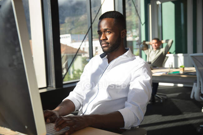 An African American businessman working in a modern office, sitting at a desk and using a computer, with his colleague sitting in the background — Stock Photo