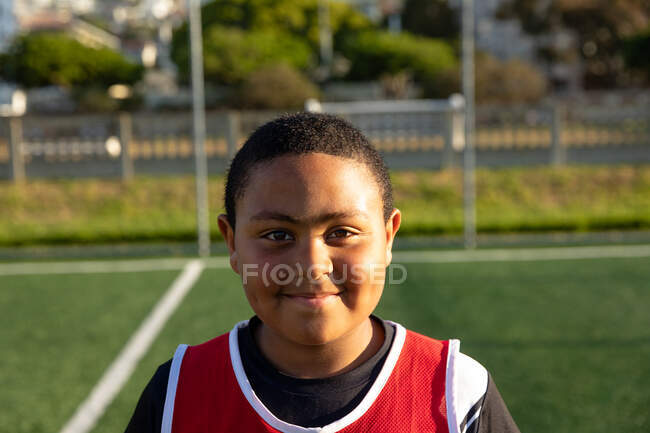 Портрет крупним планом впевнений змішаної раси хлопчик-футболіст в командній смузі, стоячи на ігровому полі на сонці, дивлячись на камеру і посміхаючись — стокове фото