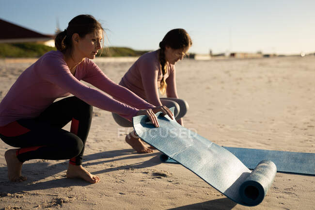 Vue latérale de deux amies caucasiennes profitant de la plage par une journée ensoleillée, préparant des tapis de yoga pour la pratique du yoga. — Photo de stock