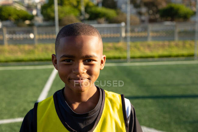 Портрет крупным планом уверенного афро-американского футболиста в командном костюме, стоящего на игровом поле на солнце, смотрящего в камеру и улыбающегося — стоковое фото