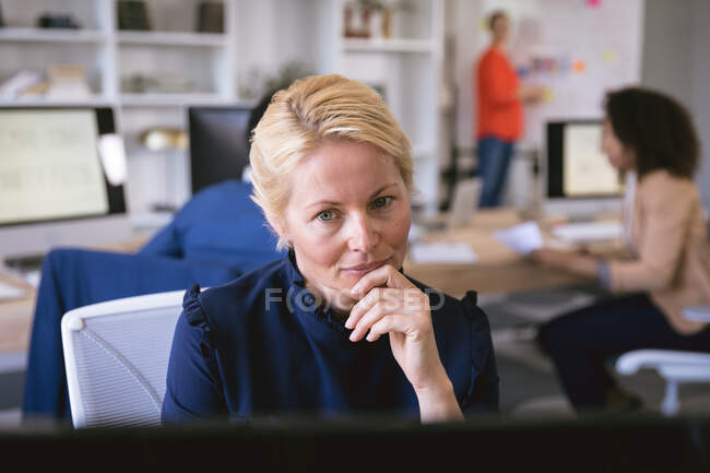 Eine kaukasische Geschäftsfrau, die in einem modernen Büro arbeitet, am Schreibtisch sitzt und einen Computer benutzt, während ihre Geschäftskollegen im Hintergrund arbeiten — Stockfoto