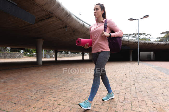 Вид спереди на белокурую женщину, идущую на тренировку в облачный день, гуляющую в городском парке с спортивной сумкой и ковриком для йоги — стоковое фото