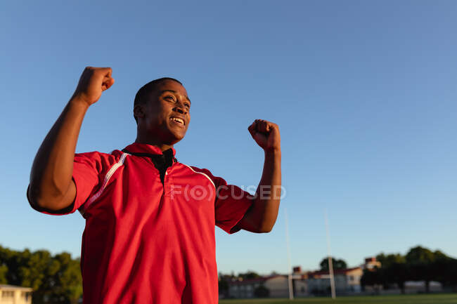 Низкий угол обзора игрока в регби юноши смешанной расы в красной команде, стоящего на игровом поле, приветствующего и поднимающего руки в честь победы во время матча по регби — стоковое фото