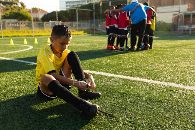 Side view of a mixed race boy soccer player sitting on a playing field in the sun putting on his football boots during a training session, with his teammates listening to their coach in the background — Stock Photo