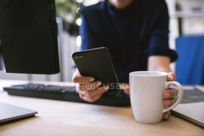 Seção intermediária close-up de empresária trabalhando em um escritório moderno, sentado em uma mesa se comunicando usando um smartphone e segurando uma caneca de café — Fotografia de Stock