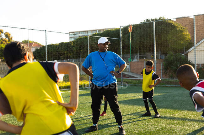 Frontansicht eines gemischten männlichen Fußballtrainers, der während eines Fußballtrainings auf einem Spielfeld in der Sonne steht und eine multiethnische Gruppe von Jungen-Fußballern mit den Händen auf den Hüften bei Stretchübungen anweist. — Stockfoto