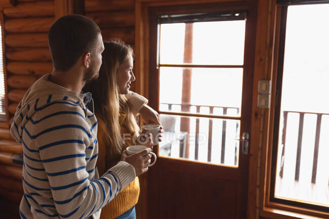 Seitenansicht eines kaukasischen Paares, das eine gute Zeit auf einer Reise in die Berge hat, in einer Hütte steht, Tassen Kaffee hält, sich umarmt, durch die Witwe blickt — Stockfoto