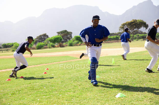 Vista laterale di un gruppo multietnico di giocatori di baseball maschi, allenamento con il loro allenatore in un campo da gioco, allenamento, corsa, in una giornata di sole — Foto stock