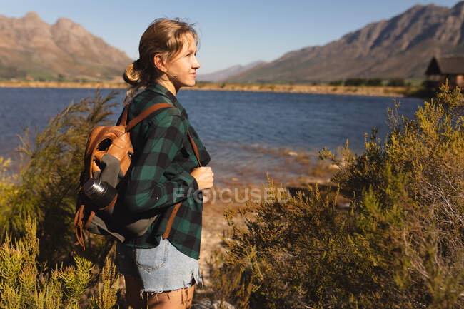 На вигляд кавказька жінка добре проводить час, подорожуючи горами, стоячи на березі озера, насолоджуючись своїм виглядом, у сонячний день. — стокове фото