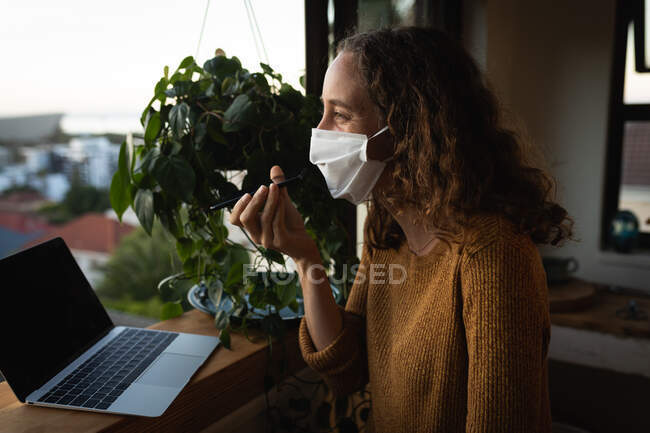 Кавказька жінка проводить час удома на самоізоляції, одягнувши маску обличчя проти коронавірусу covid19, стоячи біля вікна, розмовляючи на своєму смартфоні та за допомогою ноутбука.. — стокове фото