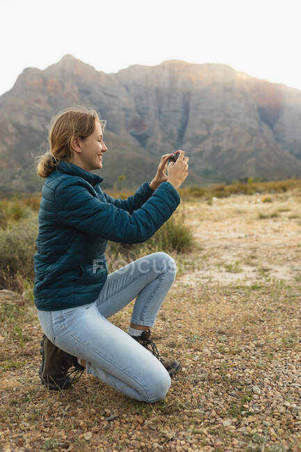 Side view of a Caucasian woman having a good time on a trip to the mountains, kneeling and taking a photo with a smartphone — Stock Photo