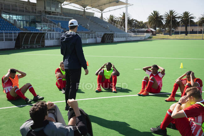 Side view of a multi-ethnic group of teenage male field hockey players and their Caucasian male field hokey coach training before a game, working out on a field hokey pitch, doing sit ups in a circle on a sunny day while the coach stands watching — Stock Photo