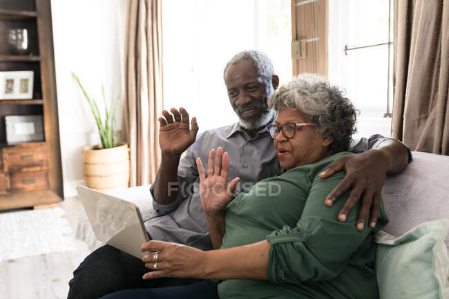 A senior African American couple spending time at home together, social distancing and self isolation in quarantine lockdown during coronavirus covid 19 epidemic, using a tablet, making a video call to friends or relatives, waving — Stock Photo
