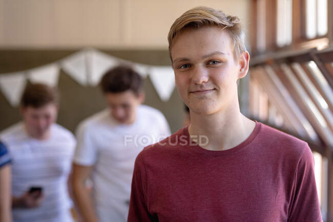 Портрет белого подростка с короткими светлыми волосами и голубыми глазами, стоящего в школьном классе и улыбающегося в камеру, с одноклассниками, разговаривающими на заднем плане — стоковое фото
