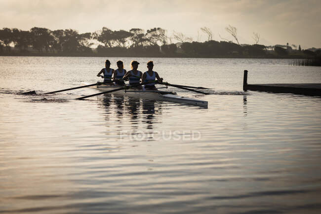 Vista frontal de un equipo de remo de cuatro mujeres caucásicas entrenando en el río, remando en una concha de carreras al amanecer, con la luz del sol reflejada en las ondas del agua en primer plano - foto de stock