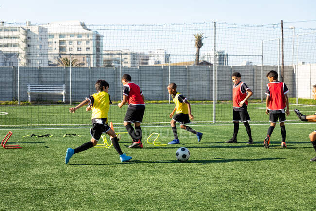 Vista lateral de un grupo de jugadores multiétnicos de fútbol masculino entrenando en un campo de juego en un día soleado, corriendo, saltando sobre obstáculos bajos y practicando habilidades de pelota. - foto de stock