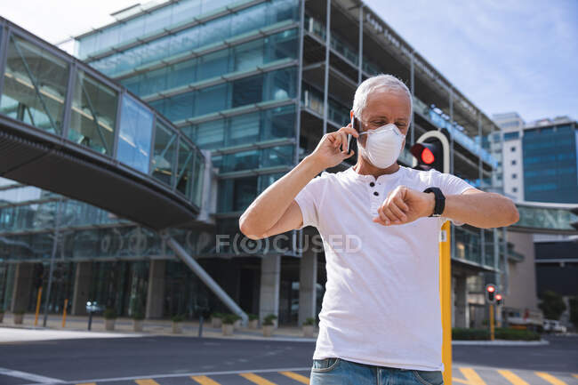 Homme caucasien âgé dans les rues de la ville pendant la journée, portant un masque facial contre le coronavirus, covid 19, à l'aide d'un smartphone et smartwatch. — Photo de stock