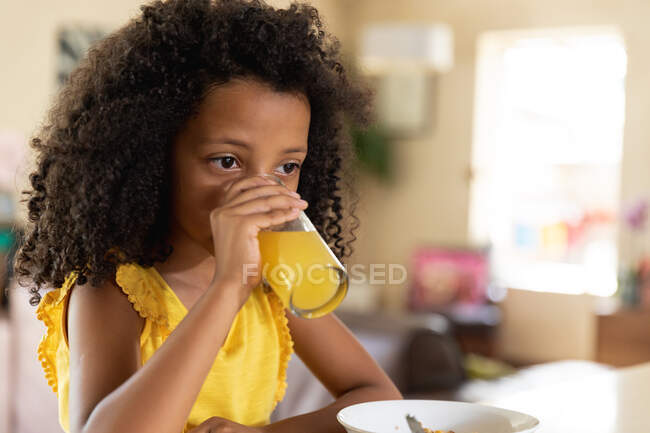 Afroamerikanisches Mädchen, soziale Distanzierung zu Hause während der Quarantäne, sitzt am Tisch und frühstückt und trinkt ein Glas Orangensaft. — Stockfoto